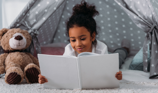 Child holding a picture book in a blanket fort with pillows and a teddy bear