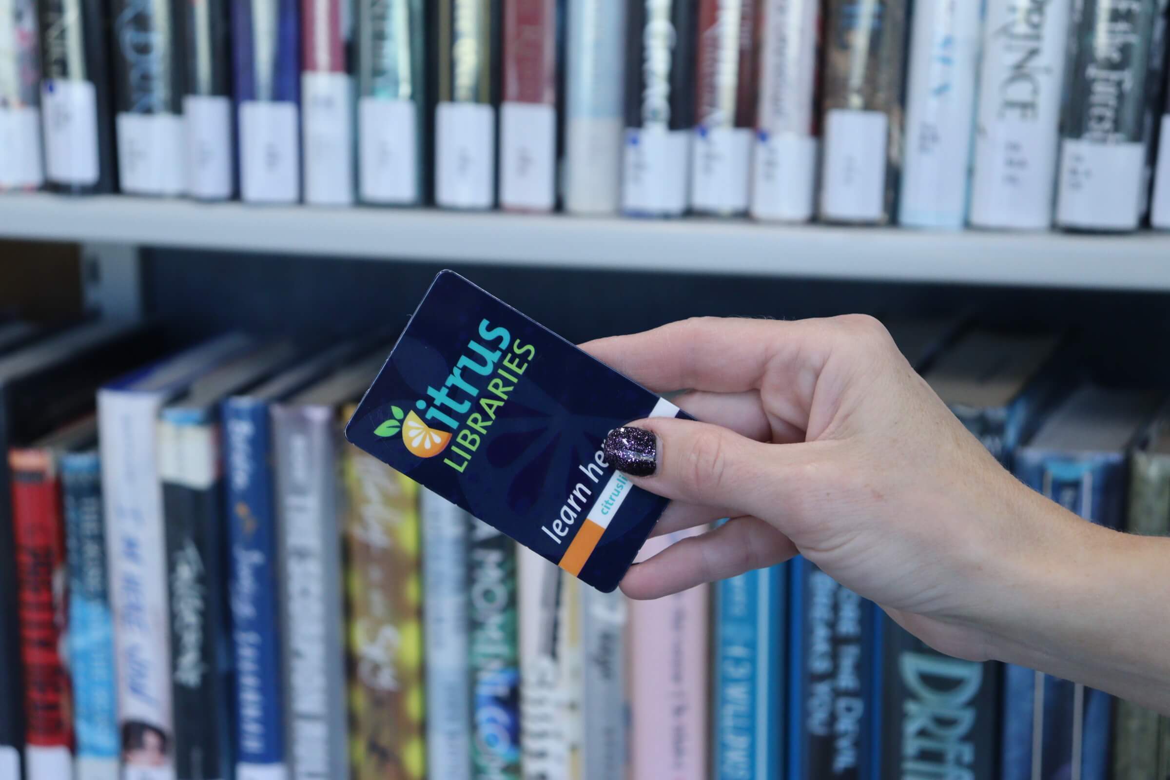 Hand holding Citrus Libraries Library Card in front of bookshelf