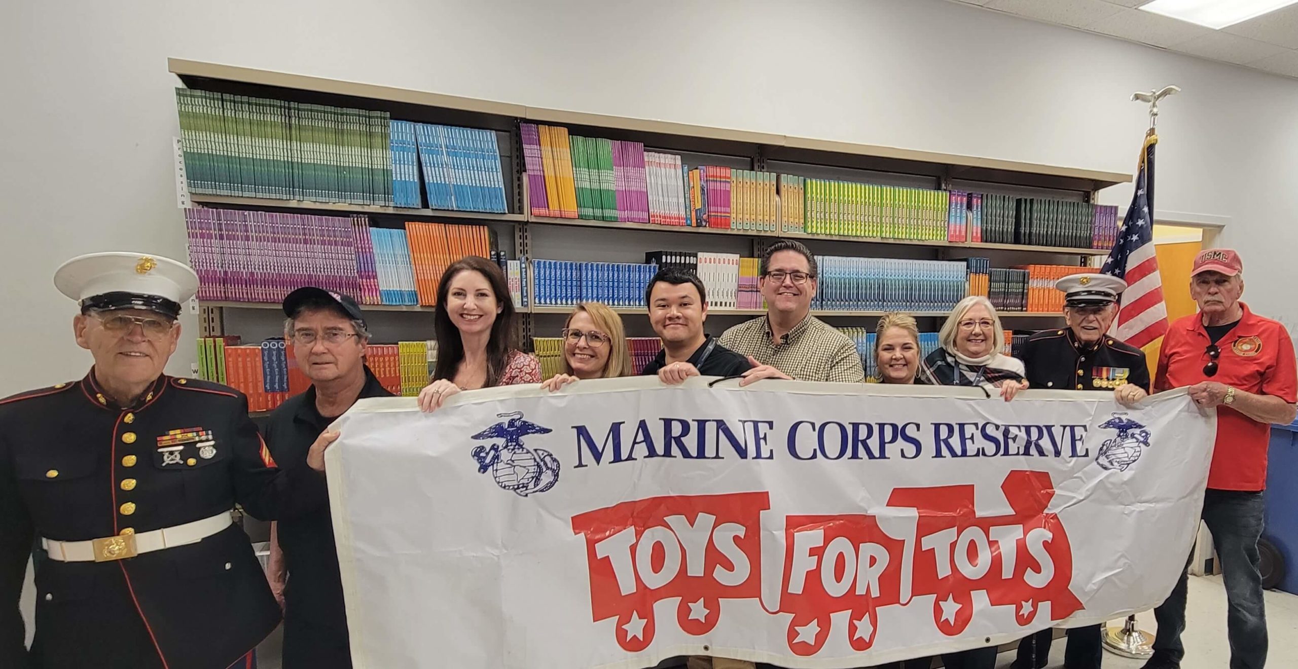 United States Marine Corps veterans and Library staff holding Toys for Tots banner in front of shelves of donated children books.