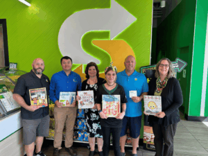  Subway Co-Owner, Scott Lambo; Library Director, Adam Chang; Region Manager, Renee Thompson; Crystal River Subway Employee; Subway Co-Owner David Lambo; Youth Librarian, Debbie Robitaille Inside a Subway Sandwich shop holding picture books in front of wall with light-up Subway logo