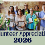 Group photo of volunteers holding award plaques with text Volunteer Appreciation 2026 on neutral green background and floral embellishments
